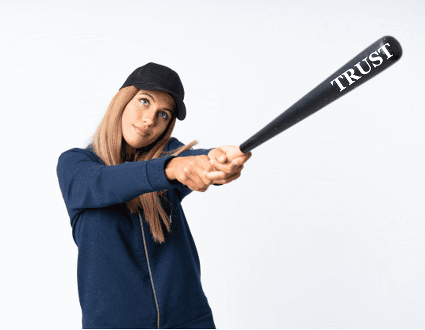 Photo of a girl wearing a dark colored baseball hat holding a black baseball bat with the word TRUST written on the bat's tip in white